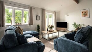A living room with a sofa set, a coffee table, a TV, and a window with curtains. - Goodwood - Modern Stay in Cotswolds with Hot Tub (South Cerney)