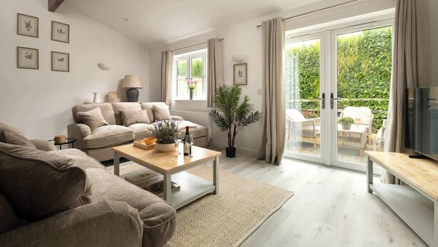 Una sala de estar moderna con un sofá, una mesa de centro y un mueble para televisión. Hay ventanas amplias con cortinas, una planta en maceta y vistas al exterior.