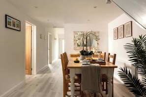 A dining area with a wooden table, chairs, a vase with flowers, and a mirror on the wall. - Epsom - Cosy Stay in the Cotswolds (Gloucestershire)