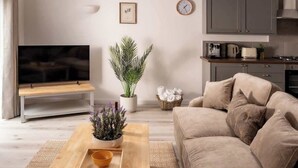 Una sala de estar moderna con un televisor de pantalla plana, una mesa de centro de madera, un sofá color beige y una cocina con armarios y estufa.