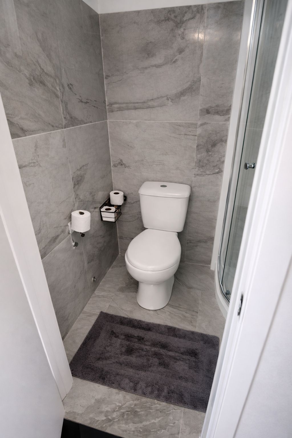 A bathroom with a toilet, marble tiles, a shower, and a dark rug.