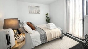 Ein modernes Schlafzimmer mit Bett, Nachttisch, Lampe und einem Fenster mit durchsichtigen Vorhängen.