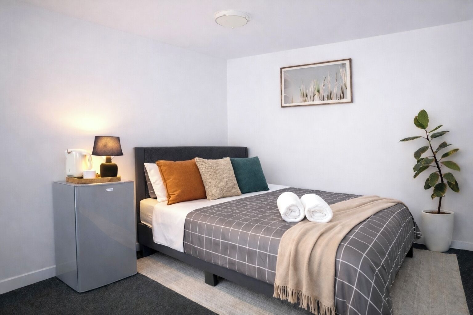 A neatly arranged hotel room with a bed, a bedside table with a lamp, a small refrigerator, and a potted plant.