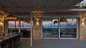 An open-plan dining area with a stone wall, large windows, and hanging lights. - Villa Arcadia (Naxos)