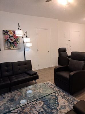 Ein Wohnzimmer mit einer schwarzen Couch, einem schwarzen Sessel, einem Glastisch und einem gemusterten Teppich. An den Wänden hängen zwei Lampen und ein Blumenbild.