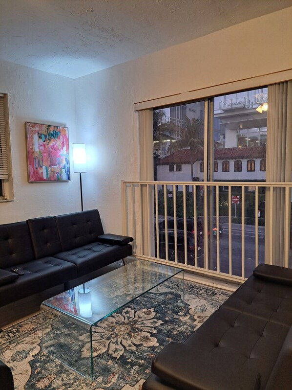 Ein Wohnzimmer mit einem schwarzen Sofa, einem Glastisch und einem Teppich mit Muster. Ein großes Fenster bietet Blick auf die Straße und die Gebäude davor.