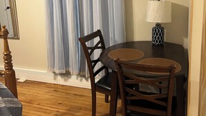 A dining area with a round table, two chairs, a lamp, and a window with blue curtains.