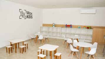 Un salón de juegos infantiles con mesas y sillas blancas, una pizarra con 'Max Kids' escrito y una estantería llena de juguetes.