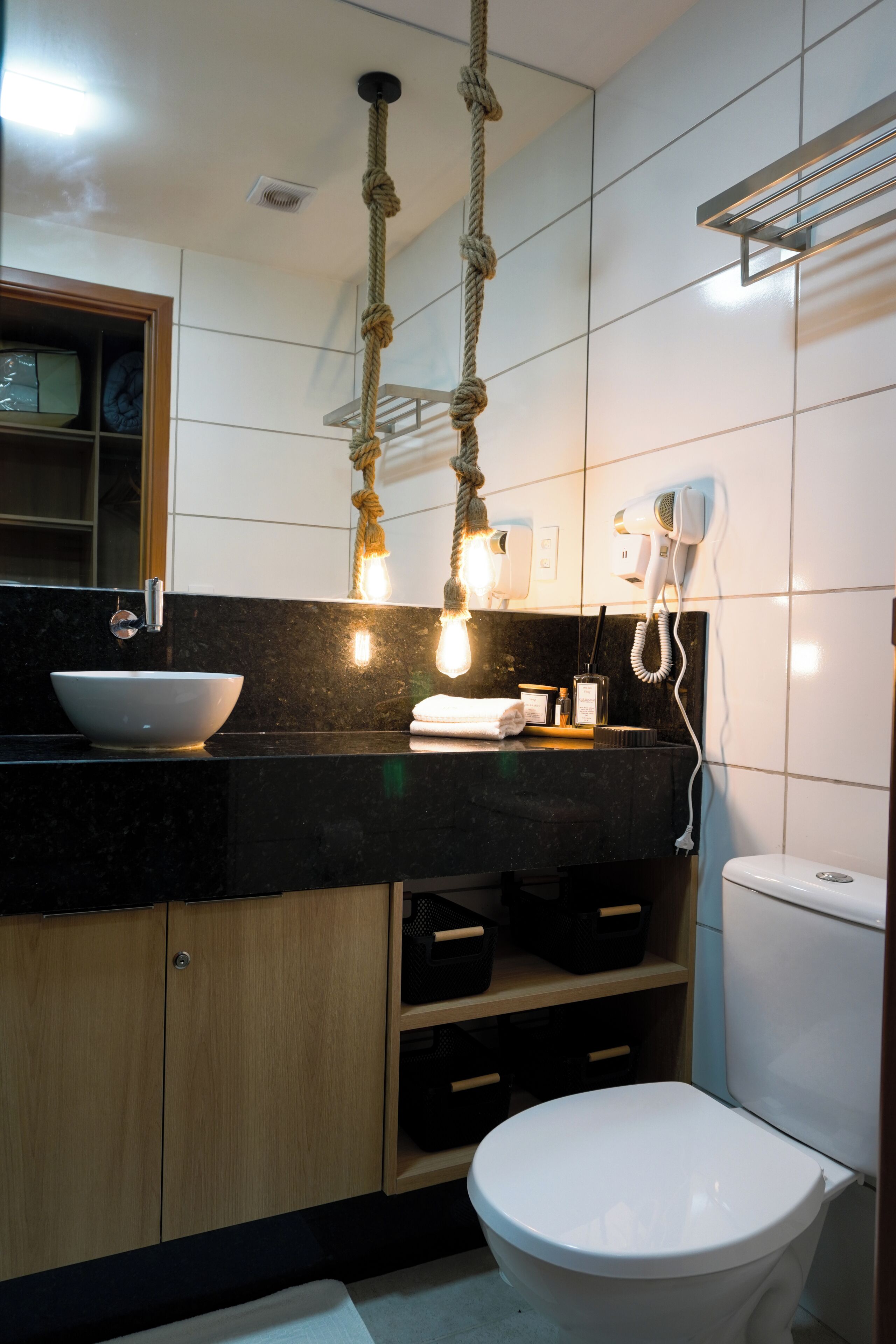 Casa de banho moderna com bancada preta, armários de madeira, sanita branca e um secador de cabelo fixado na parede.