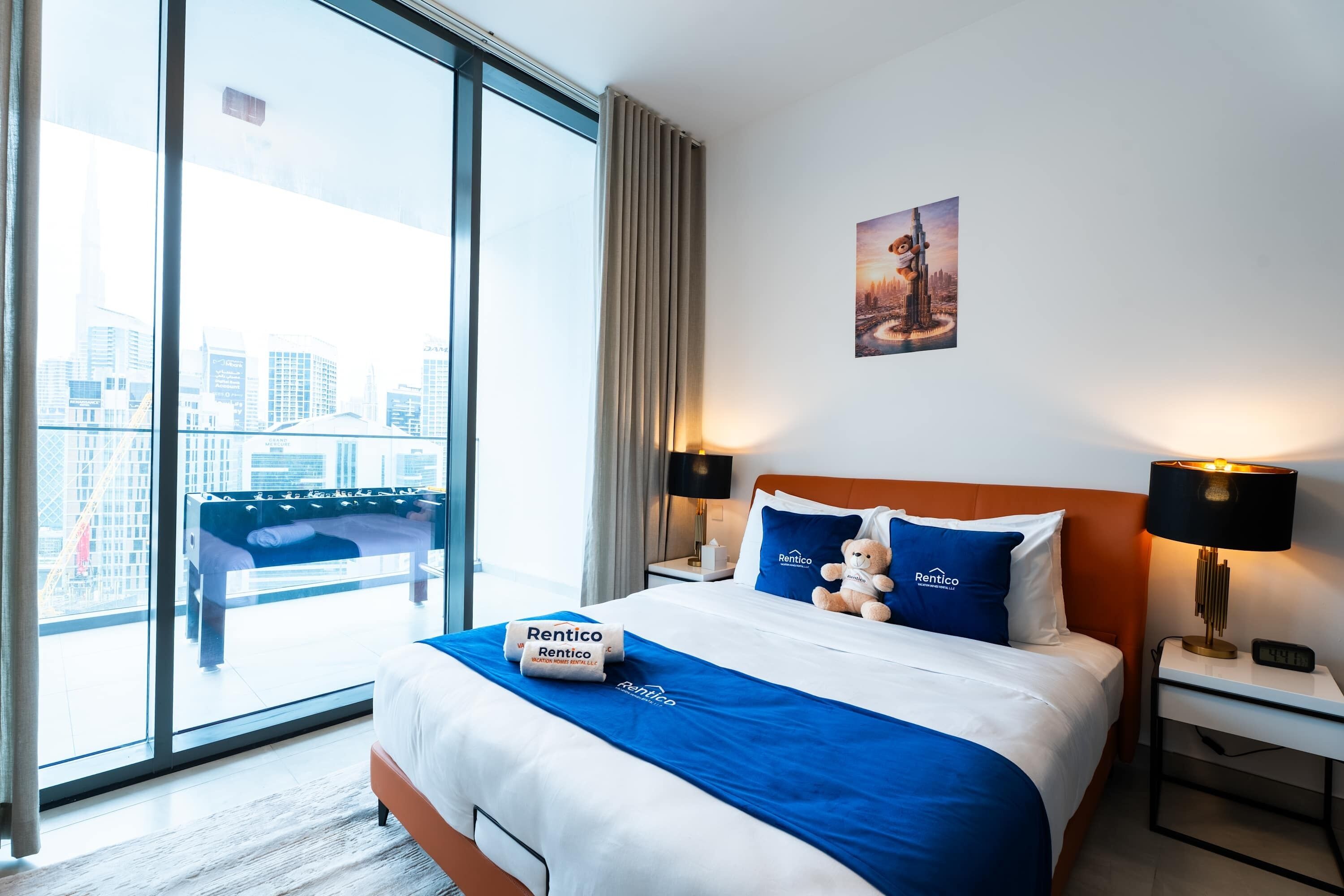 A hotel room with a large window, a bed with blue bedding, a nightstand with a lamp, and a city view.