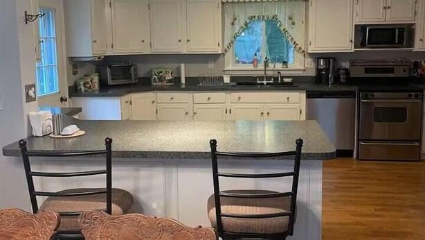 A kitchen with white cabinets, a microwave, an oven, and a dining area with two chairs and a table. - Acadia Coastal Cape 20 min to Acadia National Park (Lamoine)