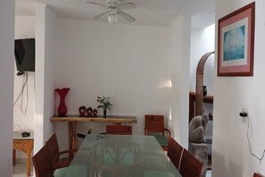 A dining area with a glass table, wooden chairs, a ceiling fan, a TV mounted on the wall, and a framed picture on the wall.