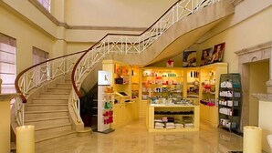 Un vestíbulo de hotel con una escalera de mármol, un letrero con forma de K, una tienda boutique y una chimenea.