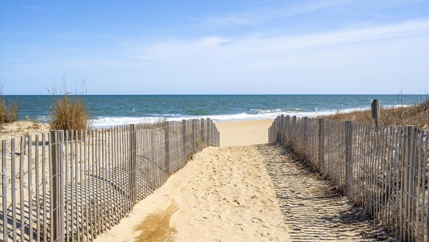 Un sentier de sable qui longe la plage et mène à l’océan, bordé de clôtures en bois et de fils de fer barbelés.