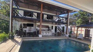 A swimming pool with a covered patio area, outdoor seating, and a kitchen visible through the open structure. - Quesada House (Tamarindo)