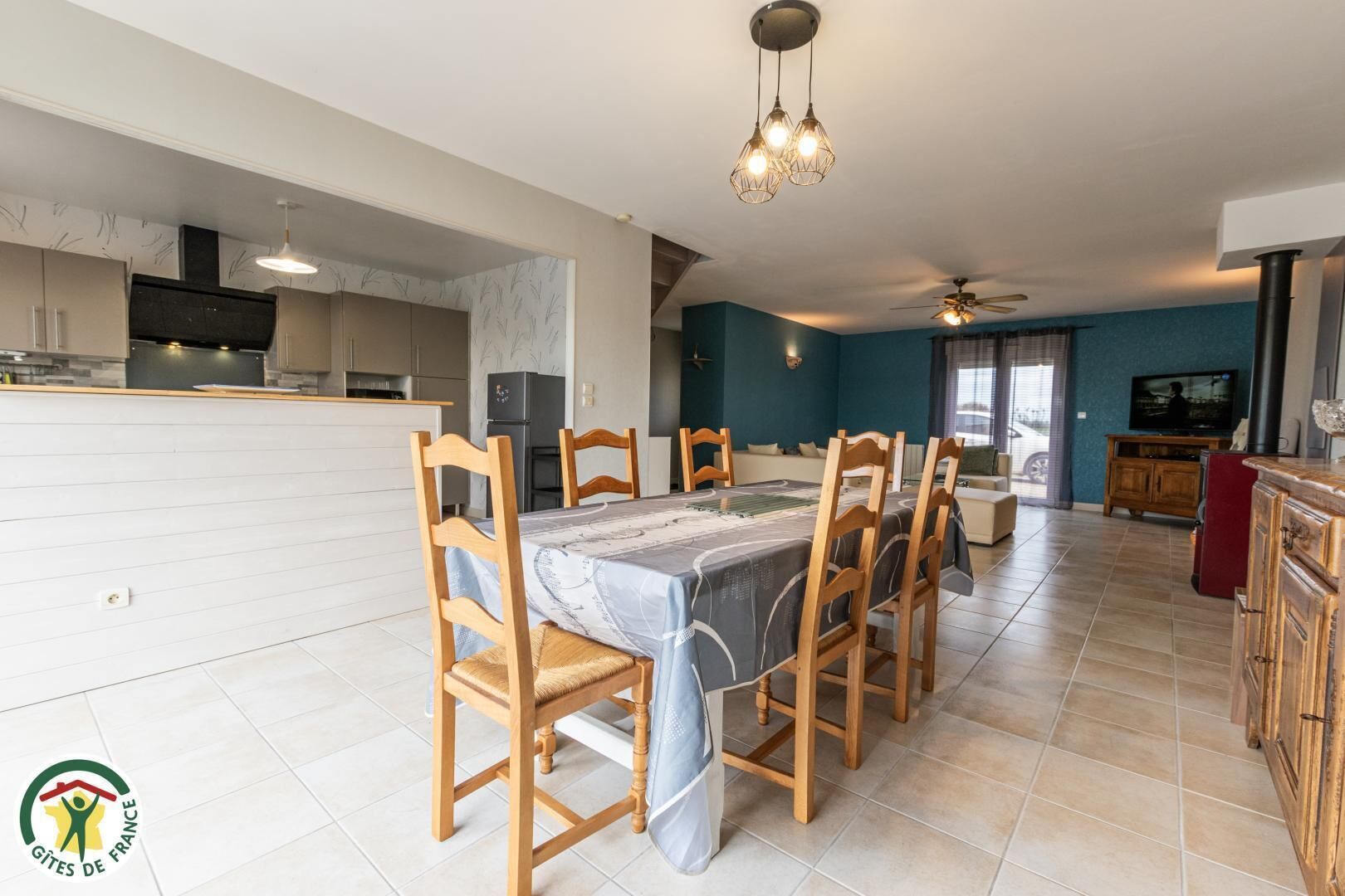Une cuisine moderne avec un coin repas, des chaises en bois, une table recouverte d’une nappe à motifs et un ventilateur de plafond.
