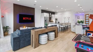 A modern living room with a large flat-screen TV displaying 'NETFLIX', a wooden bench with ottomans, a kitchen area, and a colorful arcade machine. - Oak Lawn Escape | Short Drive to Fair Park (Dallas)