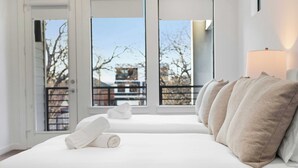 A bedroom with a large window, a bed with white linens, and a lamp on the nightstand. - Trendy Oak Lawn Retreat | Near Fair Park (Dallas)