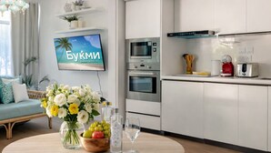 Eine moderne Küche mit weißen Schränken, einem Einbauofen, einer Mikrowelle und einer Kaffeemaschine. Ein runder Esstisch steht bereit, mit einem Blumenstrauß und einer Wasserflasche.