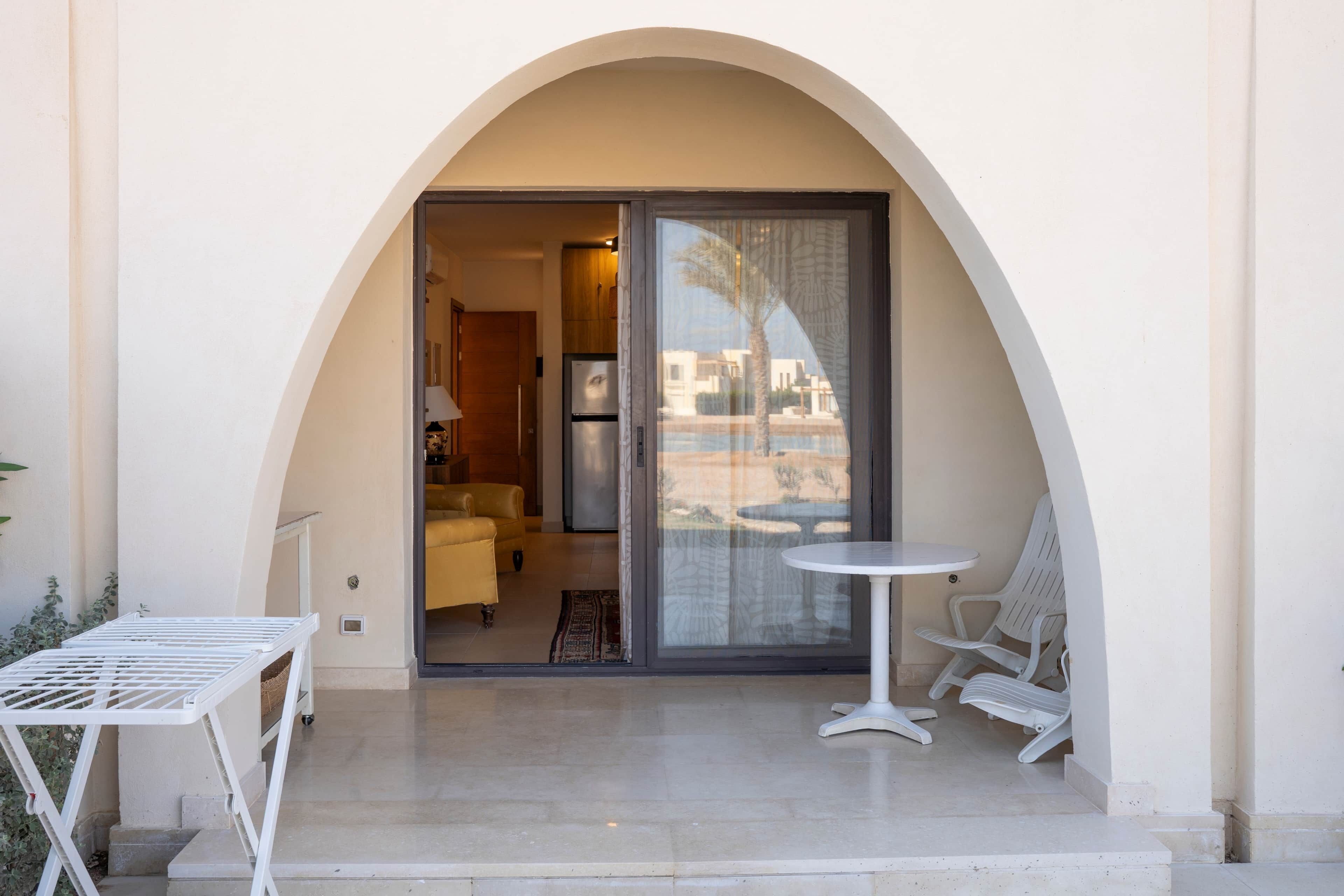Un balcón con una mesa y sillas, un ventanal corredero que da a una habitación con un sofá y una lámpara, y vistas a edificios y palmeras.