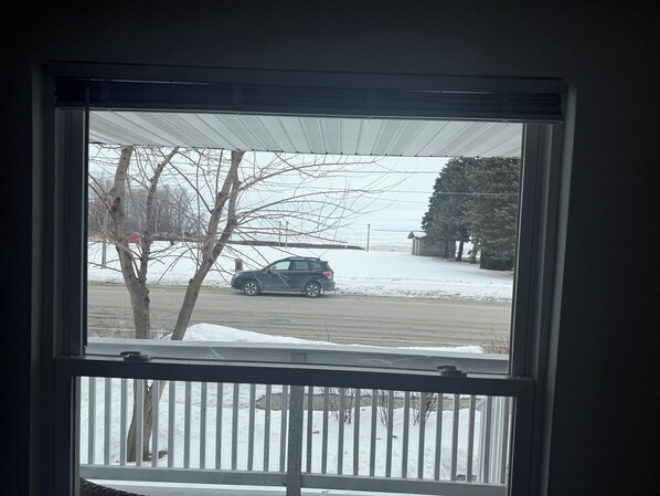 A view through a window with a metal railing, a parked car, and a snow-covered ground.