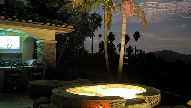 A hot tub area with a stone pool and a stone bench, surrounded by palm trees and a patio with a TV and chairs. - Luxury Wine Country Casita with Extraordinary Ridgetop Views Extensive Amenities (Fallbrook)
