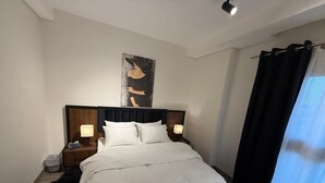 A neatly made bed with white linens, a headboard, two bedside tables with lamps, a black and white rug, and a painting on the wall. - 3-bedroom apartment in gorgeous SHIKH ZAYED with AC and comfort (giza)