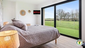 A bedroom with a large bed, a TV mounted on the wall, and a view of a grassy field through a large window.