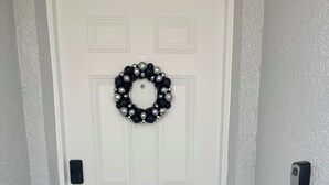 A white door with a black and silver wreath, a doorbell, and a welcome mat. - Comfort Meets Magic: Cozy Home Near Disney! (Orlando)
