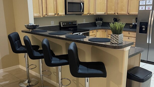 A modern kitchen with a central island, four bar stools, a microwave, a stove, and a refrigerator. - Comfort Meets Magic: Cozy Home Near Disney! (Orlando)