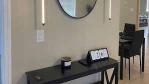 A modern living room with a round mirror, two wall-mounted lights, a black console table with a clock, and a dining area in the background. - Comfort Meets Magic: Cozy Home Near Disney! (Orlando)