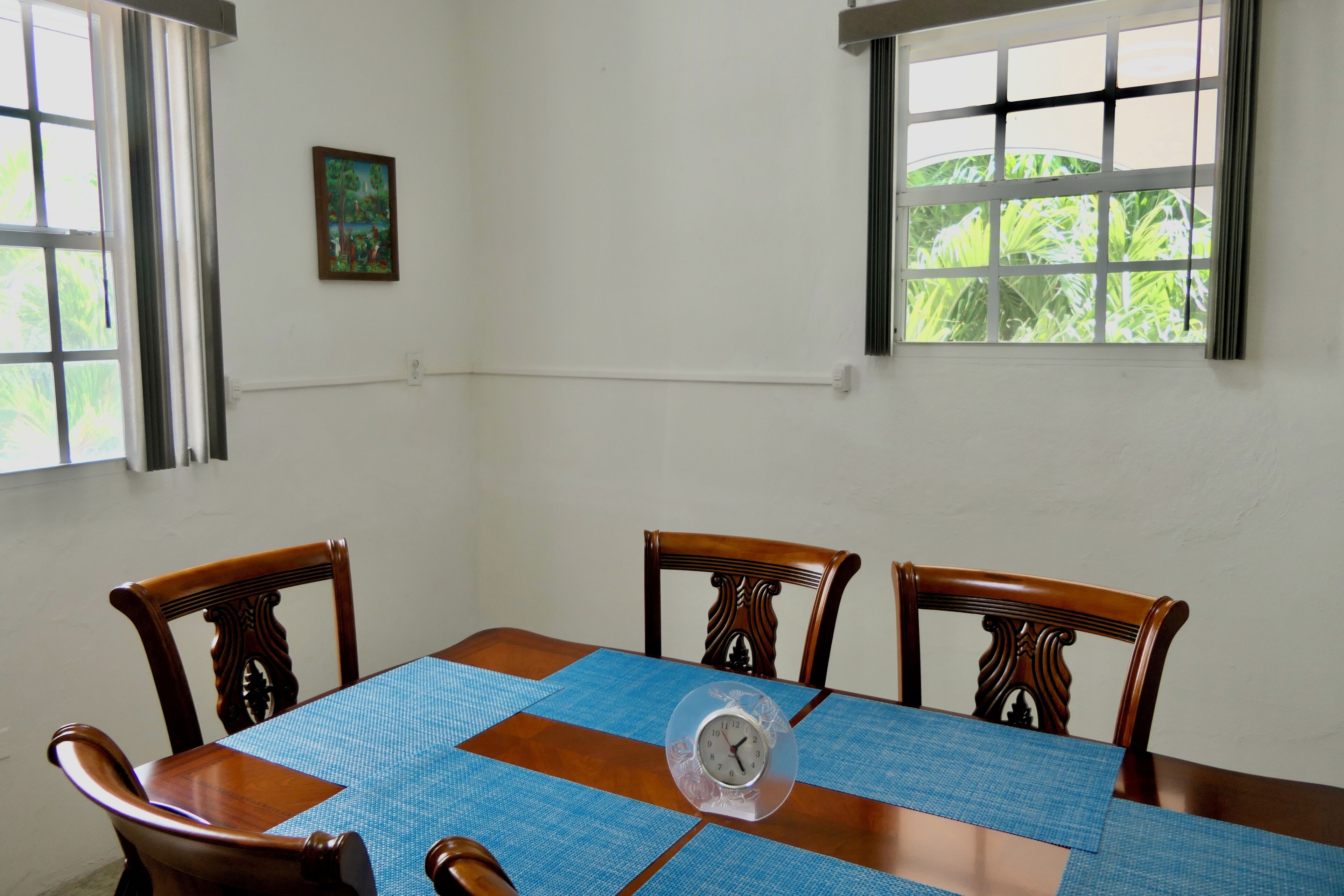 Une table à manger avec une horloge, entourée de chaises en bois, dans une pièce dotée de fenêtres et ornée d’un petit tableau au mur.