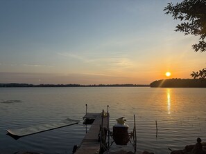 A calm lake at sunset with a dock extending into the water, a boat moored at the dock, and a distant shoreline.