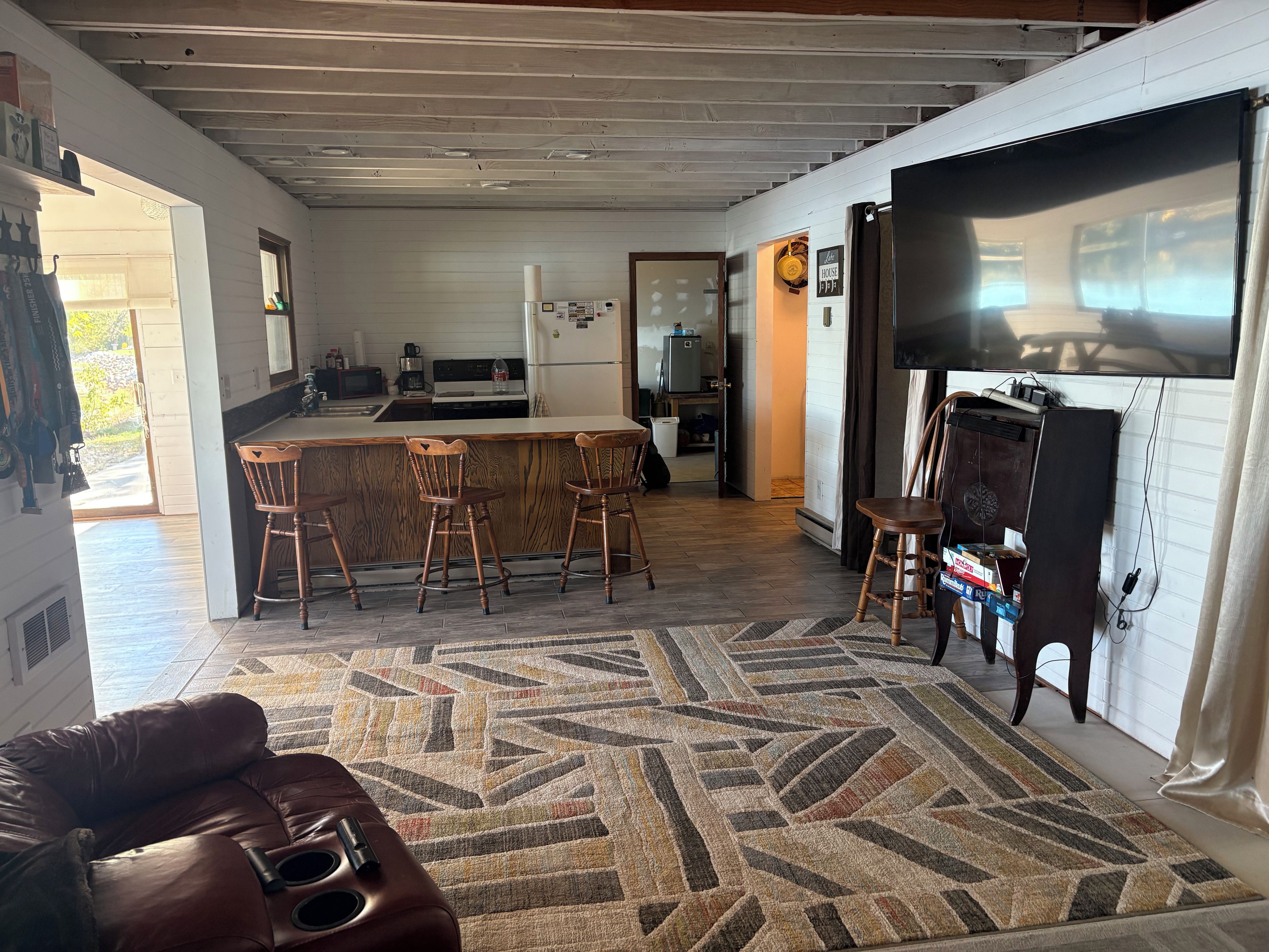 Een woonkamer met een bruine leren bank, een kamerbreed tapijt en een keukenhoek met een bar en barkrukken.