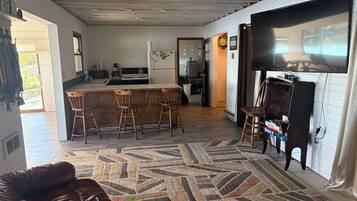 Een woonkamer met een bruine leren bank, een kamerbreed tapijt en een keukenhoek met een bar en barkrukken.
