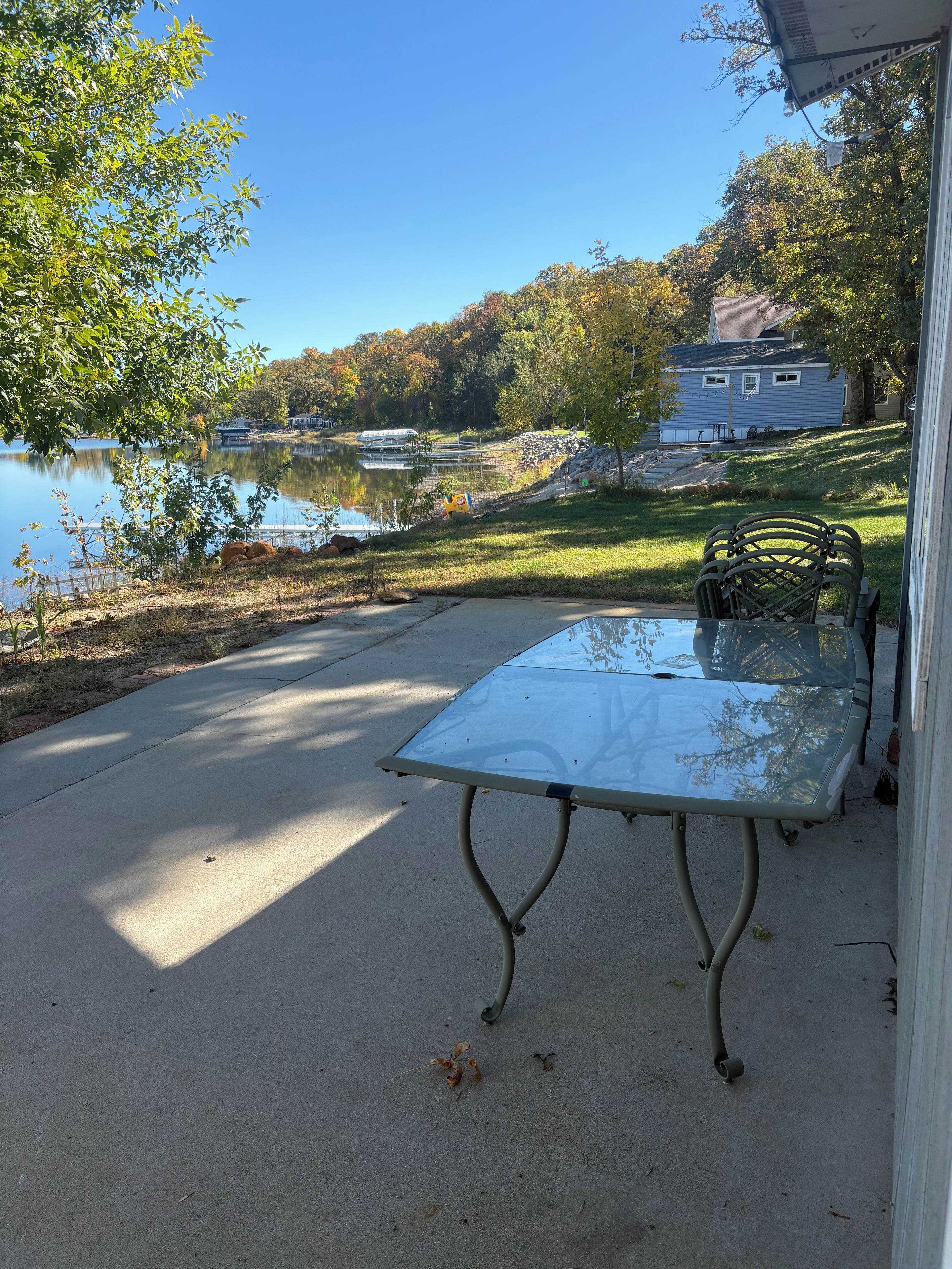 A glass-top table with metal legs on a concrete patio, overlooking a lake with docked boats and a house in the background.