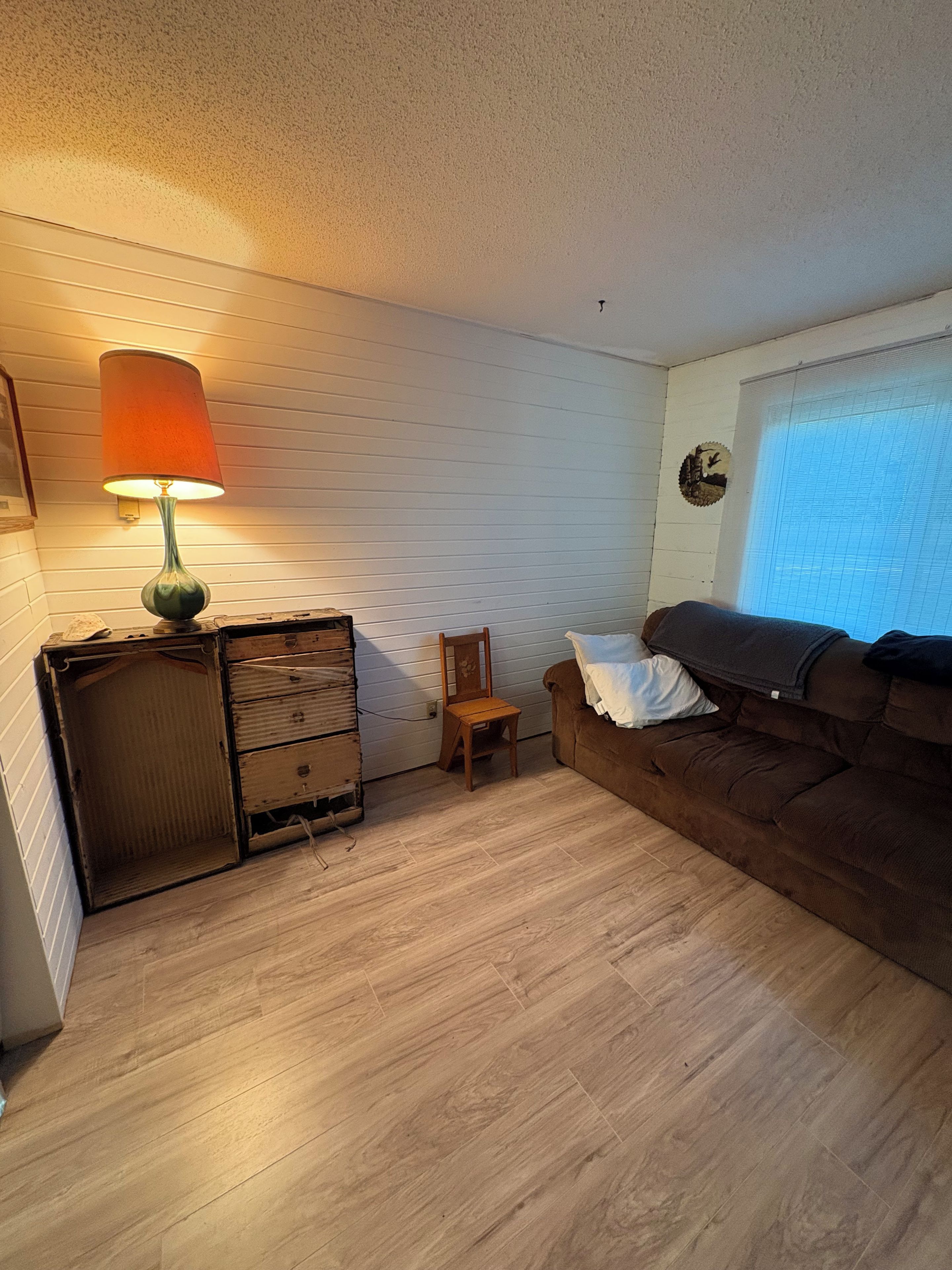 Een kamer met een houten vloer, een bruine bank, een klein houten stoeltje, een lamp en een dressoir.