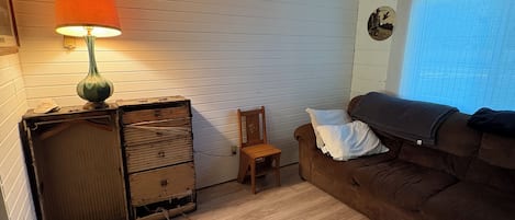 A room with a wooden floor, a brown sofa, a small wooden chair, a lamp, and a dresser.