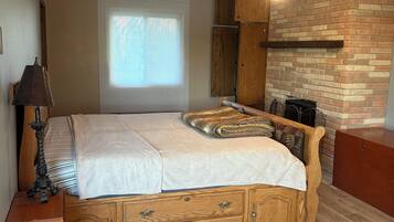 A bedroom with a bed, wooden drawers, a ceiling fan, a window with blinds, and a brick wall.