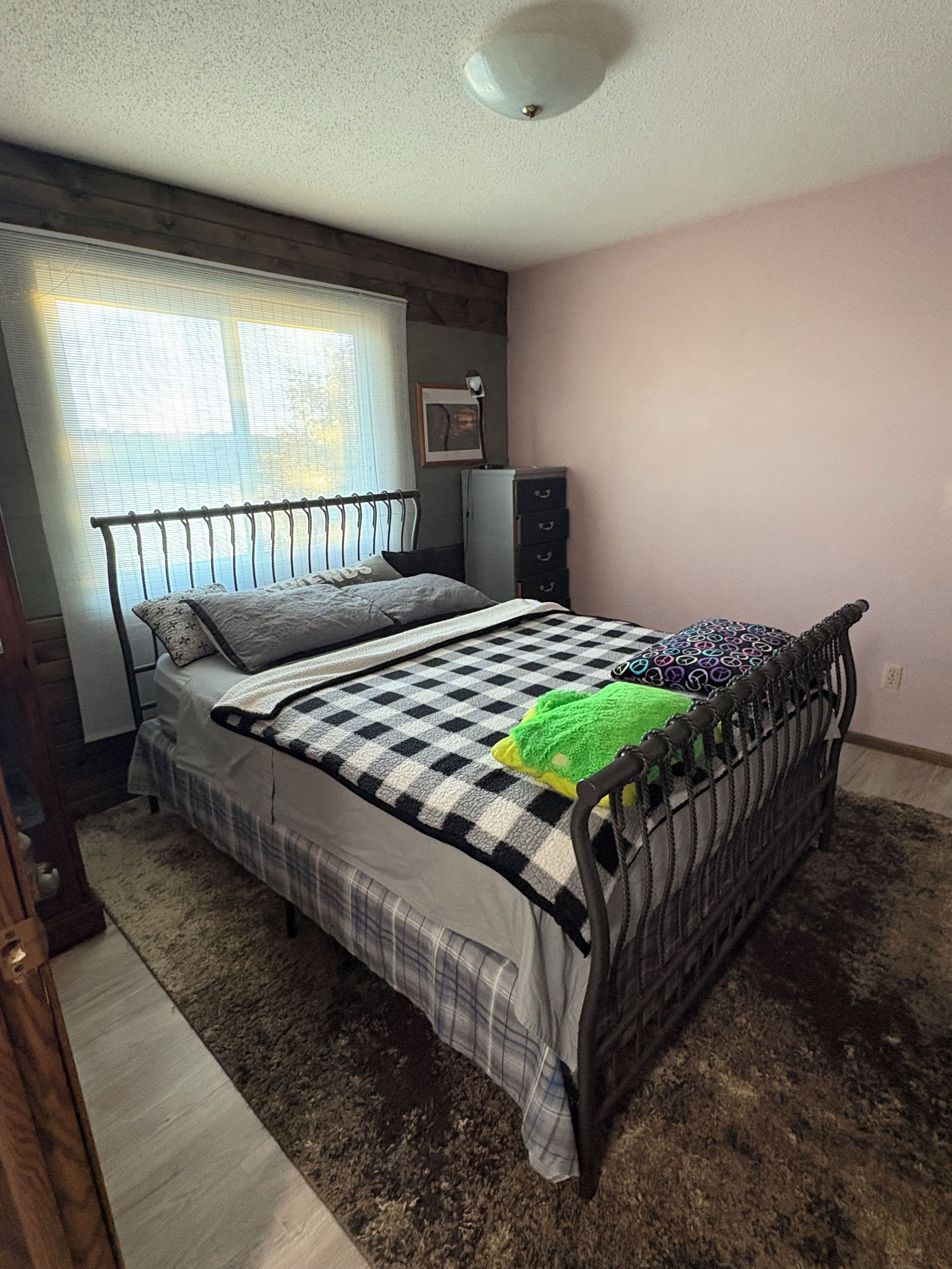 Un dormitorio con estructura de cama metálica, colcha a cuadros, manta verde, cómoda y radiador.