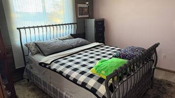 Un dormitorio con estructura de cama metálica, colcha a cuadros, manta verde, cómoda y radiador.