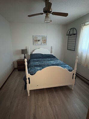 A bedroom with a bed, a ceiling fan, a nightstand, a lamp, a window with a curtain, and a wall-mounted picture.