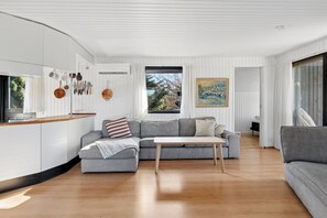 A modern living room with a grey sofa, a wooden coffee table, and a kitchen area with white cabinetry and hanging utensils. - Holiday home for 2 families by the water (Væggerløse)