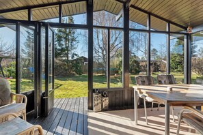 A covered outdoor area with a table and chairs, large windows, and a view of a yard. - Holiday home for 2 families by the water (Væggerløse)