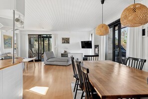 A spacious dining area with a large wooden table and chairs, a television mounted on the wall, and a large window providing natural light. - Holiday home for 2 families by the water (Væggerløse)