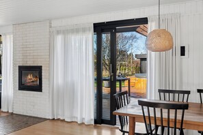 A room with a fireplace, a dining table with chairs, a large window with curtains, and a view of an outdoor deck. - Holiday home for 2 families by the water (Væggerløse)