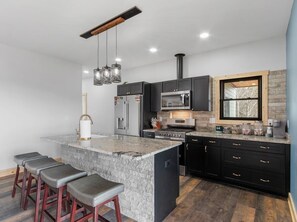 A modern kitchen with a large island, black cabinetry, stainless steel appliances, and a wood-burning stove. - Mountain Reflections | Mtn View+Sunsets & Fire Pit (Newland)