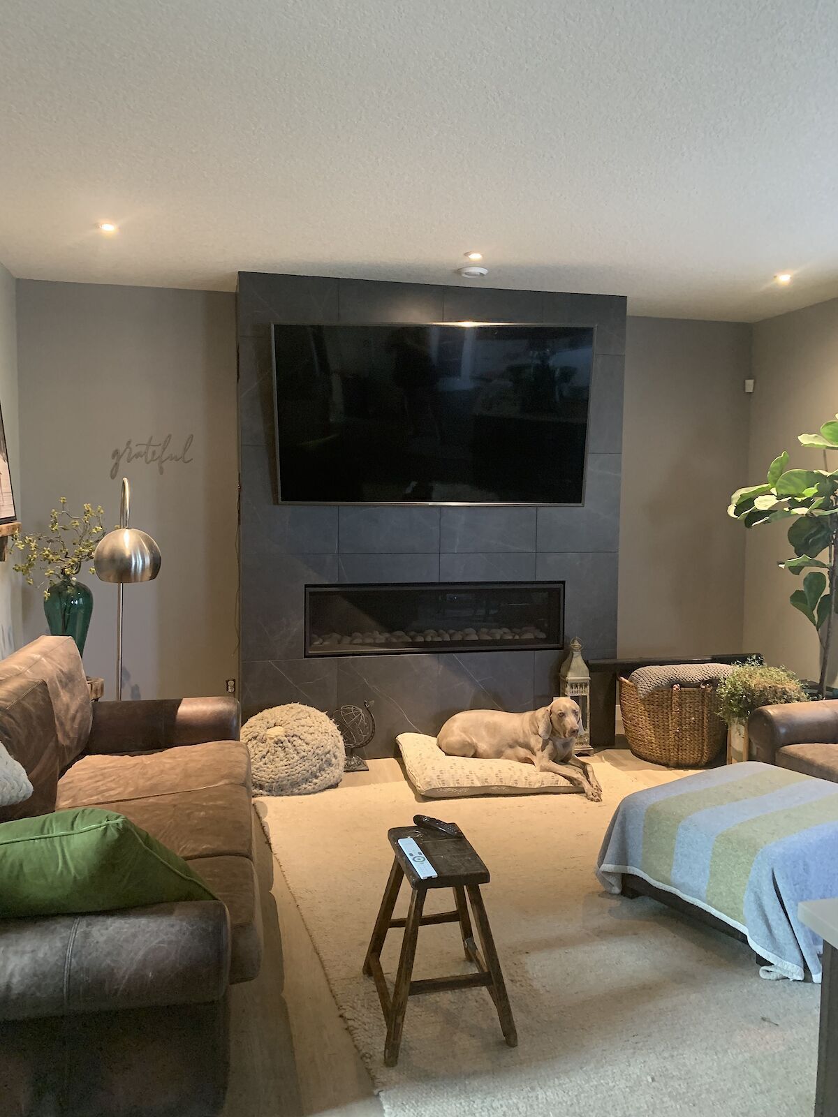 A modern living room with a flat-screen TV mounted on the wall, a fireplace below, leather sofas, a small wooden stool, and a potted plant.