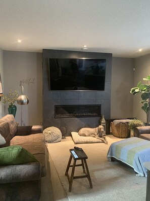 A modern living room with a flat-screen TV mounted on the wall, a fireplace below, leather sofas, a small wooden stool, and a potted plant. - Stylish Calgary Haven in Cougar Ridge – Quiet Street, Close to Parks & C‑Train (Calgary)
