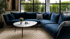 A modern living room with a blue sectional sofa, a white coffee table, and a large window with a view of greenery and a building. - Lovely holiday home near beach with sauna (Otterup)